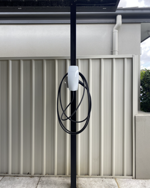 White Tesla Wall Connector with glowing green status light mounted on a slim black post in a covered carport, black cable looped around the unit