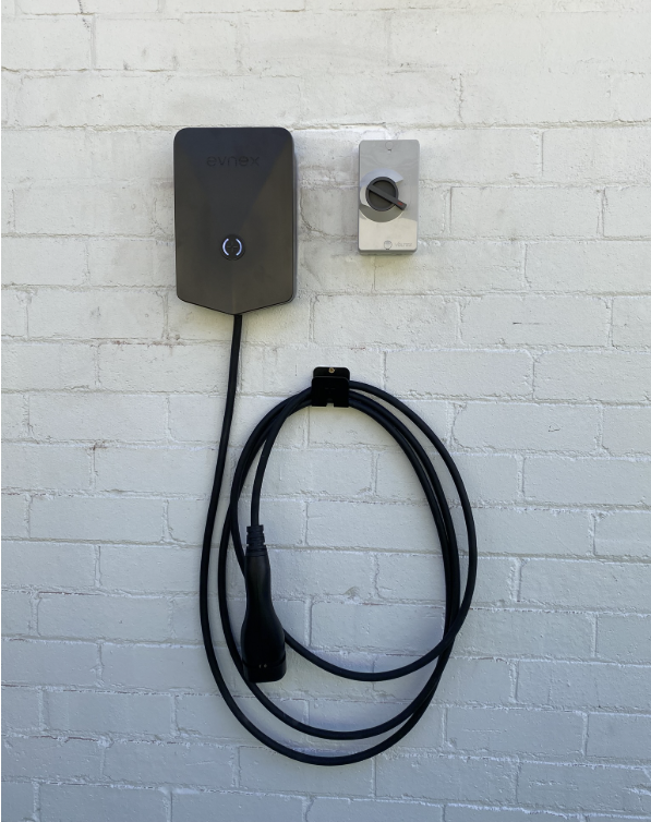 Black Evnex EV charger with glowing status light mounted on a white wall beside a grey rotary isolator, with the charging cable coiled on a hook below