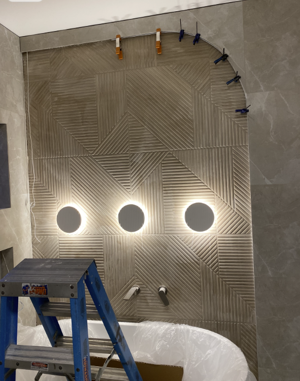 Bathroom feature wall of fluted beige tiles with three round backlit wall lights above a freestanding bath