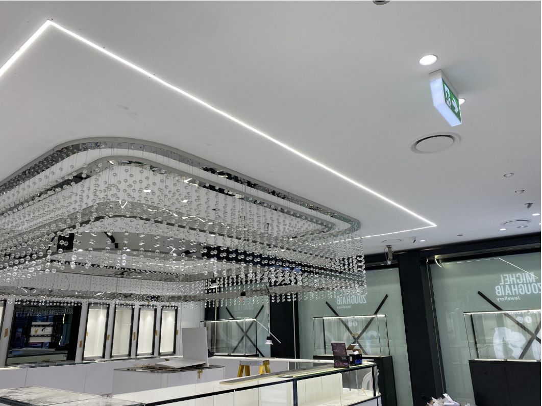Jewellery store interior during fit-out with a recessed linear LED ceiling channel, crystal bead chandelier, recessed downlights and an emergency exit sign