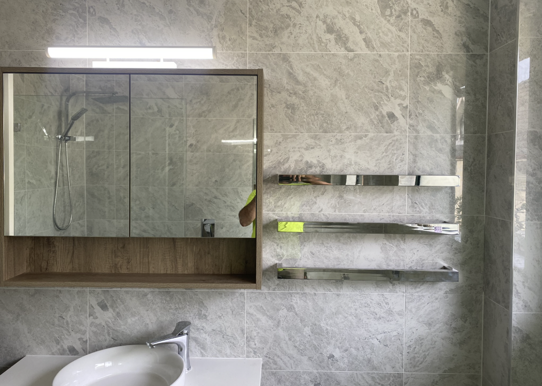 Three chrome heated towel rails on a grey marble-look tiled bathroom wall beside a mirrored vanity cabinet and round basin