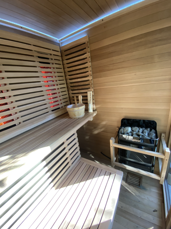 Wooden sauna with overhead LED strip, glowing infrared panels behind slatted backrests and a Harvia electric heater with stones inside a wooden guard rail