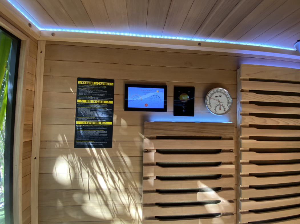 Sauna wall with touchscreen control panel, digital thermostat display and Harvia thermo-hygrometer under blue LED ceiling lighting
