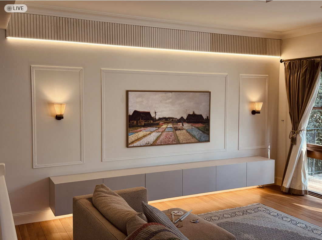 Living room with warm LED strip lighting under the upper wall panel, underglow beneath a floating media unit and two pleated-shade wall sconces